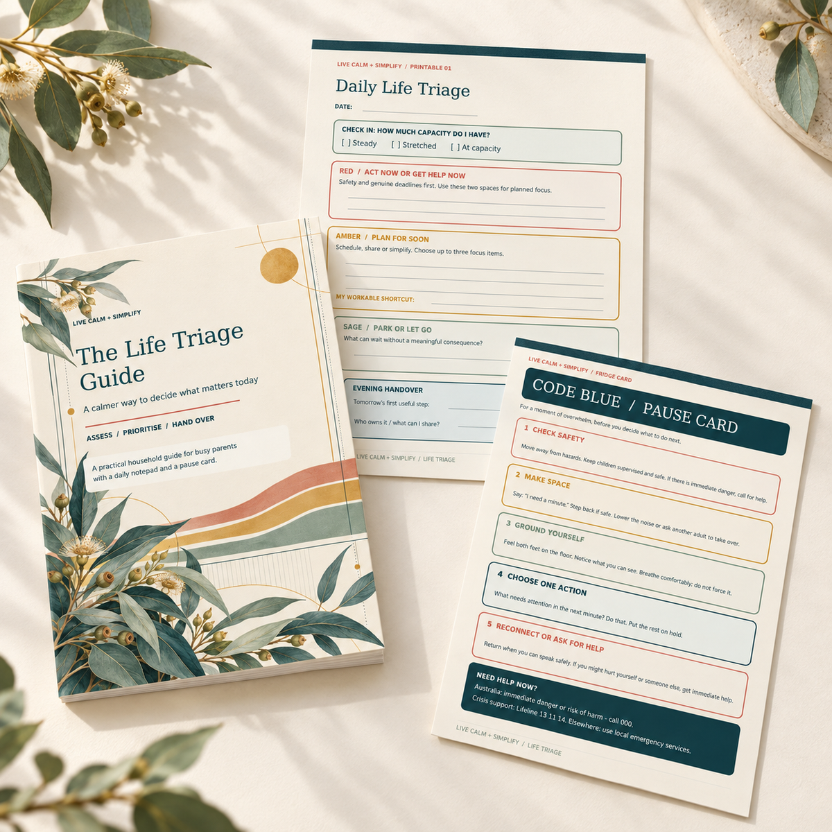 A set of mental health first-aid guide cards and a notebook titled 'The Life Triage Guide' arranged on a light fabric background with decorative leaves. The items include a notebook cover, a daily life triage sheet, and a code blue / pause card.
